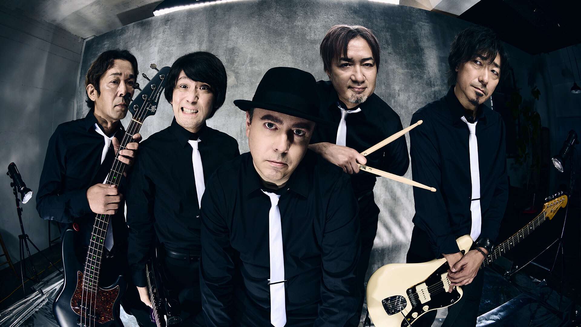 Ein Foto der Bandmitglieder der Primals. Sie tragen schwarze Hemden sowie weiße Krawatten und einige Mitglieder halten Gitarren und Schlagzeugstöcke. Im Hintergrund ist eine graue Betonwand.
