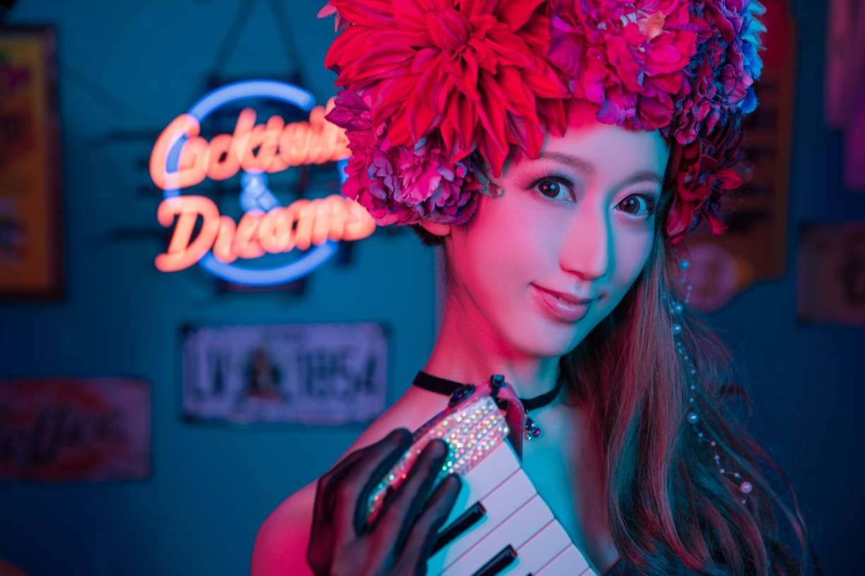 A vibrant, colorful photo of Keiko with flowers in her hair. A neon sign and various wall-mounted adornments hang in the background.