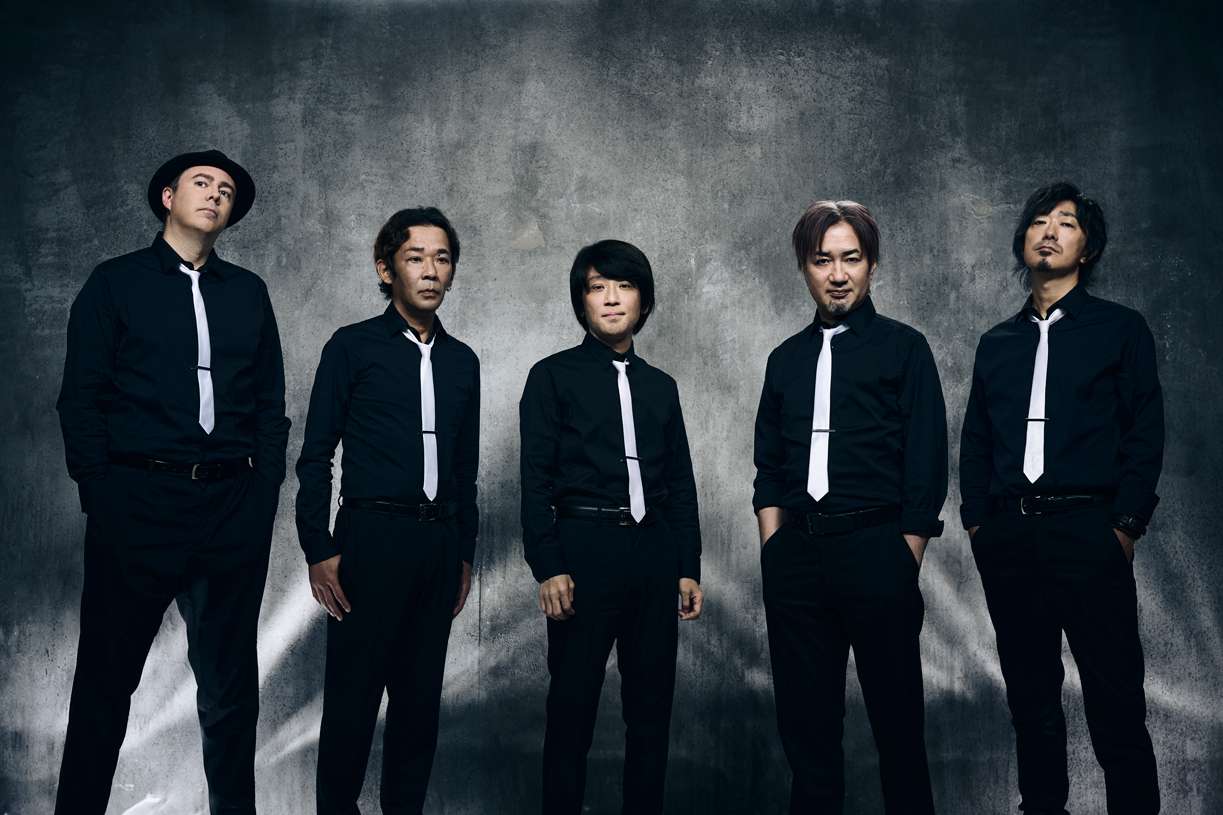 The members of the Primals band standing in a row, wearing black slacks and dress shirts with white ties.